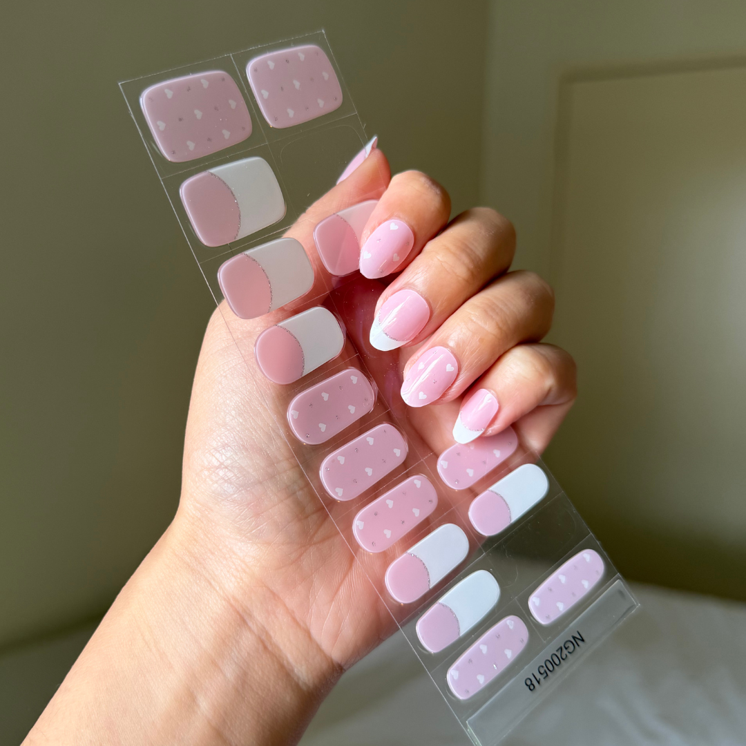 Hand holding a sheet of gellae  pink nail stickers with love hearts and white french tipon  a neutral background
